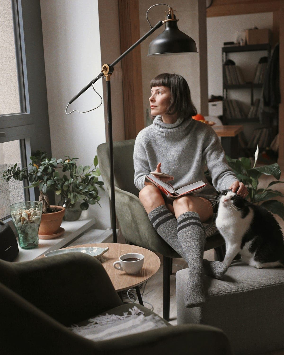 A woman reading in an armchair by a window while her cat sits on the arm beside her